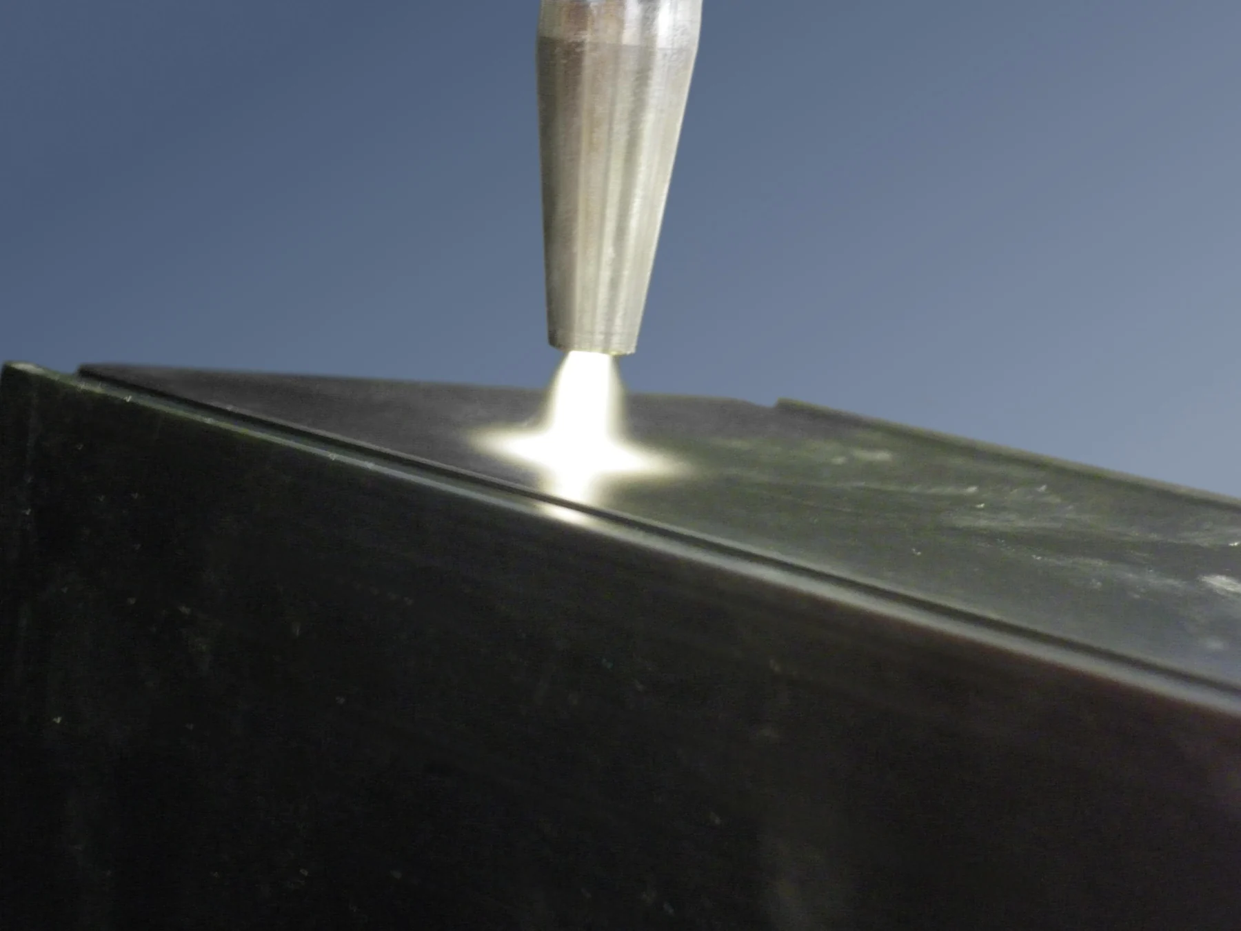 A narrow plasma plume leaves a metal nozzle above a dark flat surface.