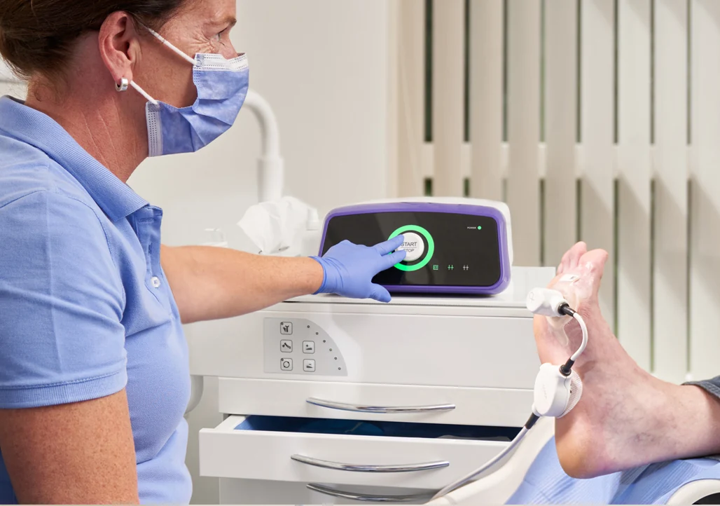 A masked practitioner operates a cold-plasma device beside a patient's supported foot.