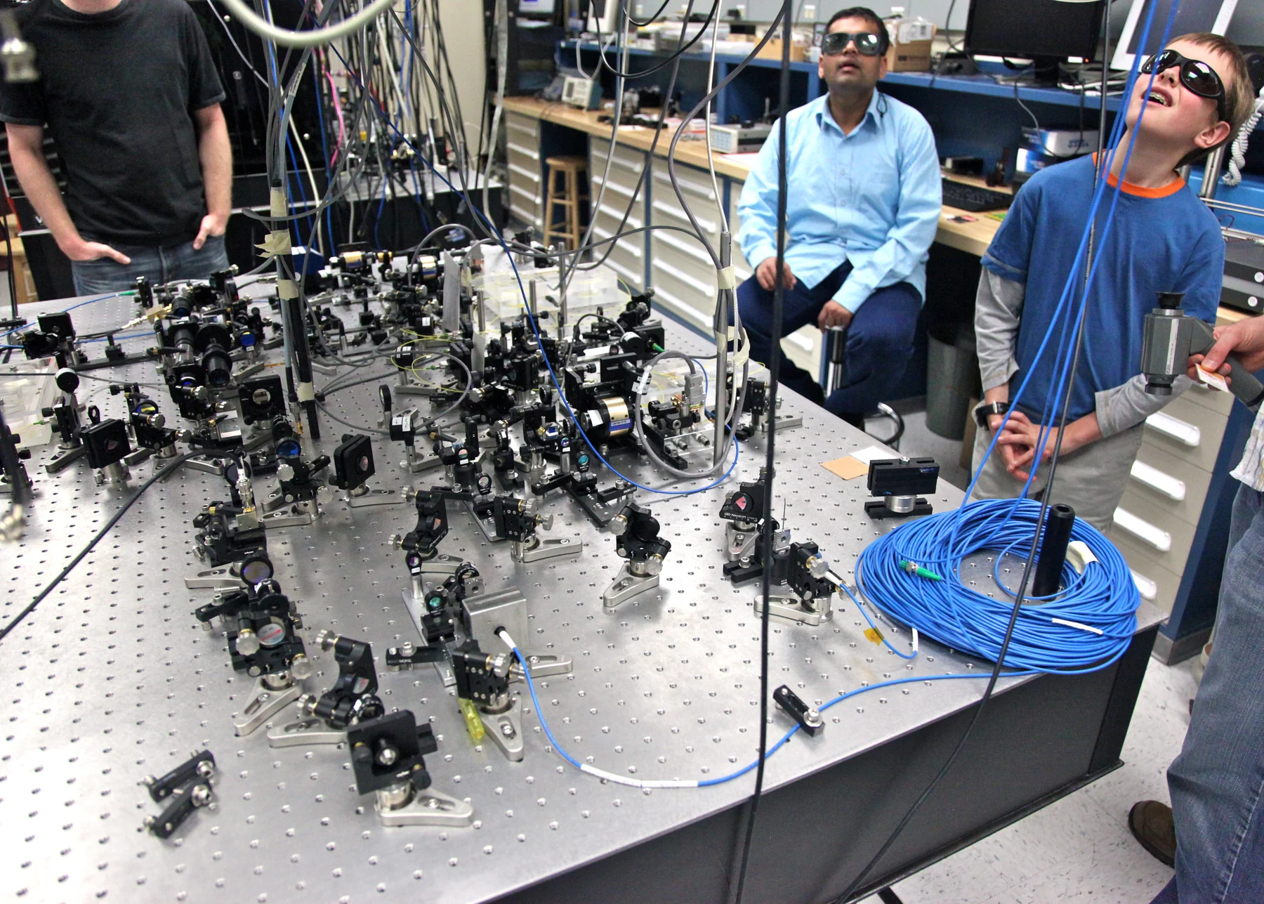 Researchers stand around an optical table crowded with mirrors, mounts, and cables.