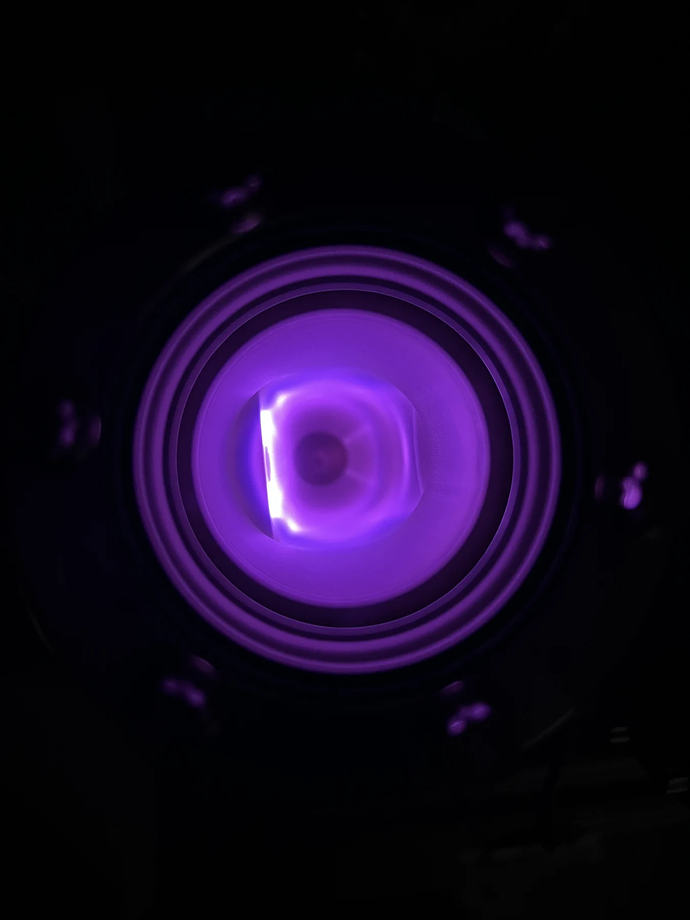 A violet plasma glows through a round port in a dark laboratory chamber.