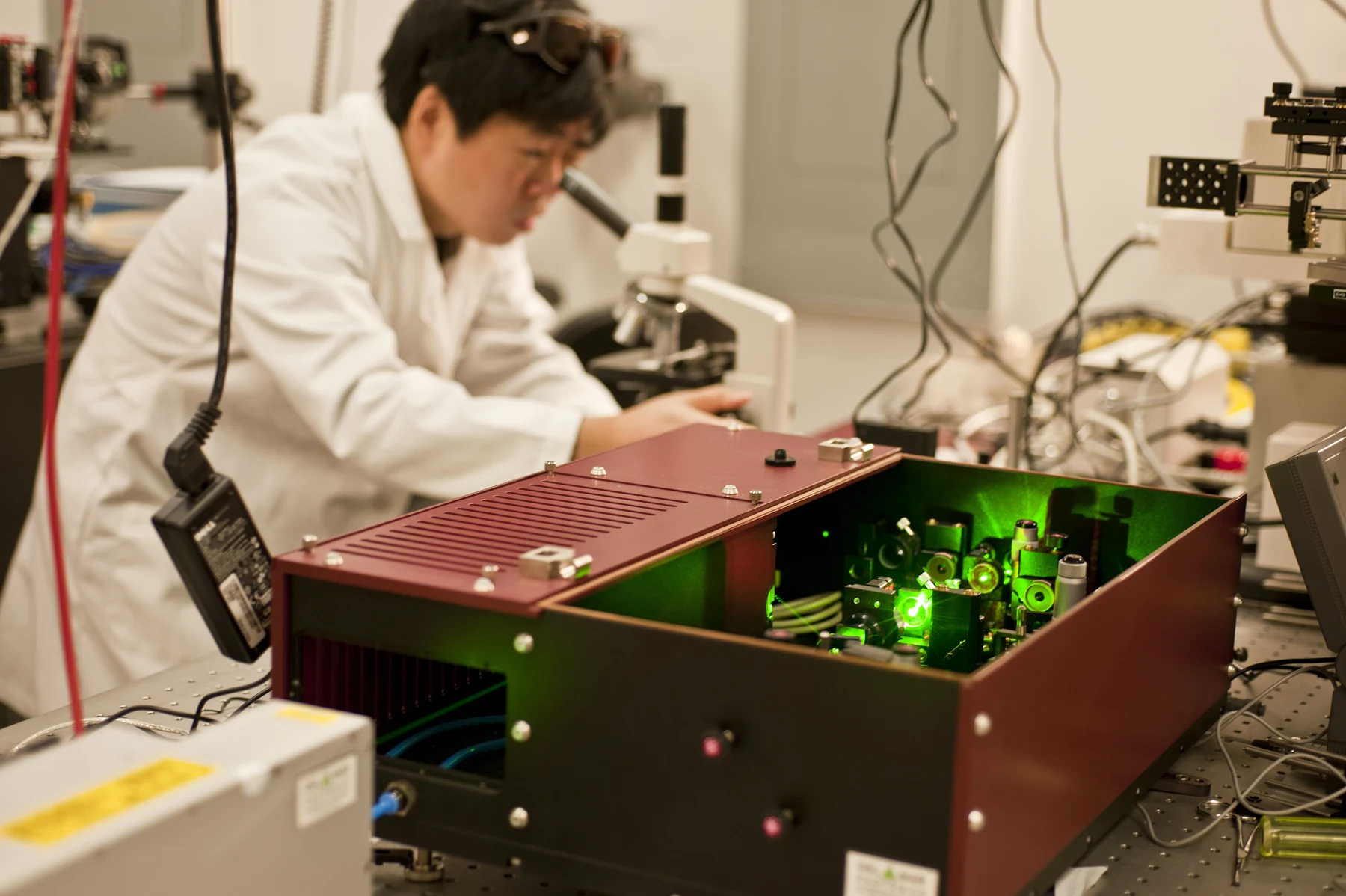 A scientist wearing protective glasses adjusts an instrument with green light inside an optics laboratory.