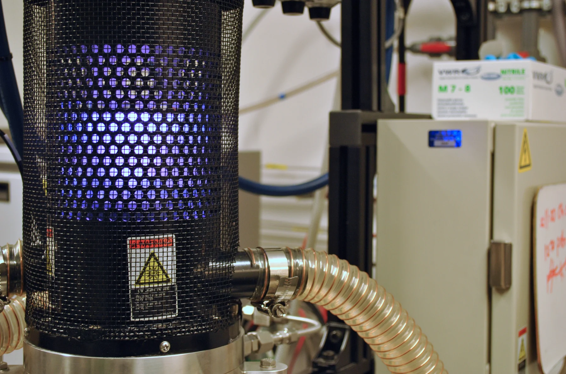 A cylindrical deposition chamber glows violet behind a perforated safety enclosure.