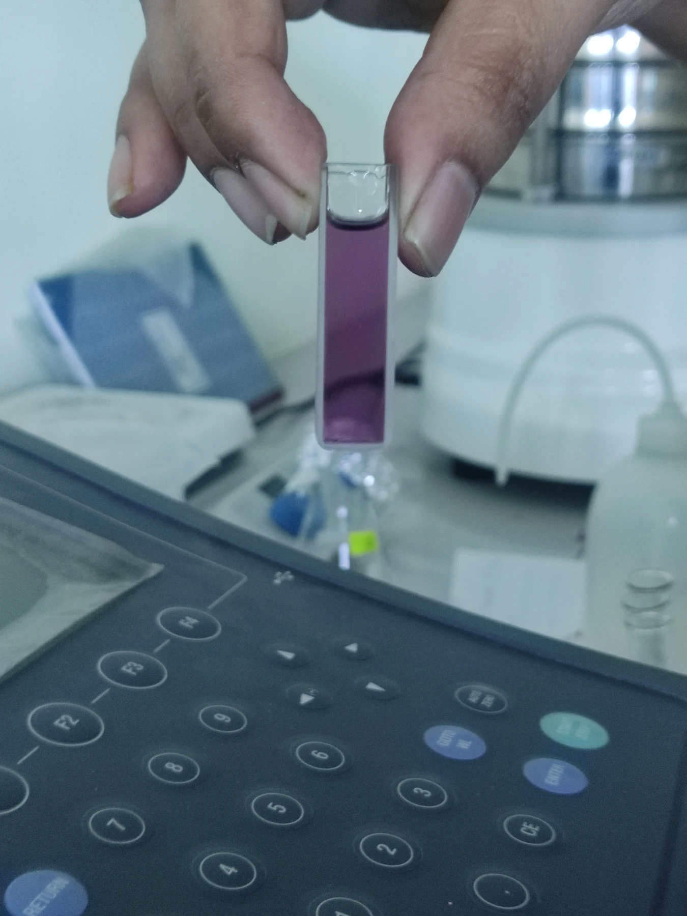 A hand holds a violet liquid sample in a cuvette above a laboratory spectrometer.